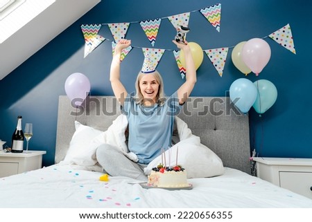 Emotional happy woman in party cap with raised arms, celebrating win in video games. Girl with console sits on a bed in a decorated bedroom. Celebrate birthday at home, pajamas party. Selective focus
