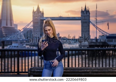 A pretty, blonde city woman looks at her smart phone during sunset time in front of the urban skyline of London