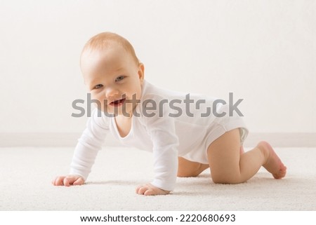 
Happy smiling adorable baby boy in white bodysuit crawling on knee and arms on light beige home carpet. Posing and looking at camera. 5 to 6 months old infant development. Royalty-Free Stock Photo #2220680693