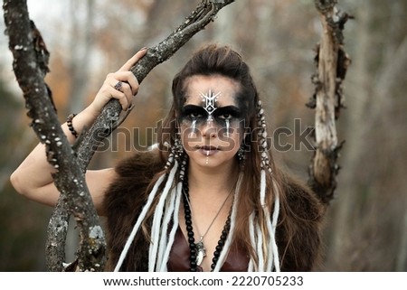 Autumn portrait of a female viking wearing face paint, braids, leather and fur. She is standing in the forest.  Royalty-Free Stock Photo #2220705233