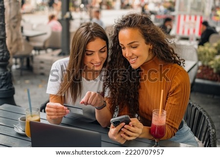 Two girls are sitting in the cafe, one is looking something at her smart phone while the other is checking information on her credit card. Royalty-Free Stock Photo #2220707957
