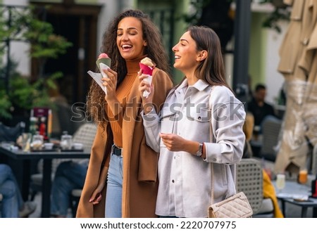 Two girls are strolling, talking and eating cone ice cream, and having much fun in the town. Royalty-Free Stock Photo #2220707975