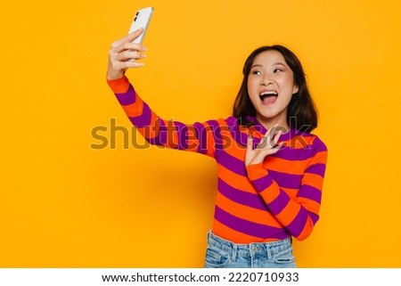 Asian young woman in stripped shirt laughing and taking selfie photo isolated over yellow wall