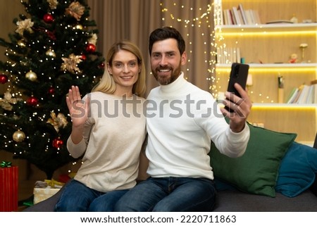 Portrait of happy christmas couple with smartphone to record online greeting video, husband and wife looking at camera celebrating new year sitting on sofa in living room.