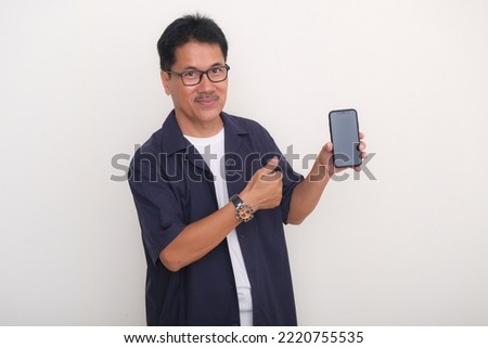 A middle-age Asian man wearing glasses shows a cell phone he is holding in his left hand; smiling, happy, friendly expression. Royalty-Free Stock Photo #2220755535