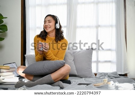 Beautiful and cheerful young Asian woman wearing headphones, holding a coffee mug, enjoys listening to music while relaxing in her modern minimal living room. home lifestyle concept