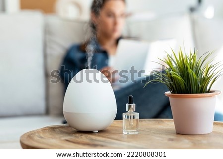 Shot of essential oil aroma diffuser humidifier diffusing water articles in the air while woman reading a book sitting on coach. Royalty-Free Stock Photo #2220808301