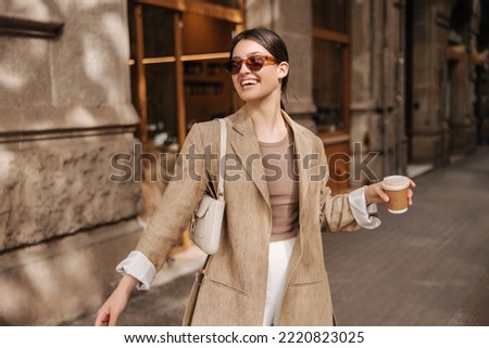Happy young caucasian woman in good mood walks with coffee around city during day. Brunette wears sunglasses, jacket and bag. Real emotions concept