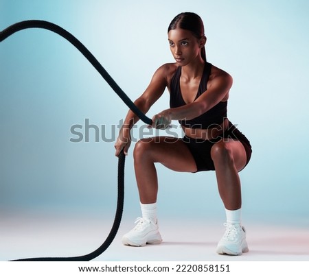 Battle rope, fitness and woman in studio for exercise, strength training and cardio workout burning calories. Wellness, focus and healthy girl in a squat pose moving ropes with a blue background Royalty-Free Stock Photo #2220858151