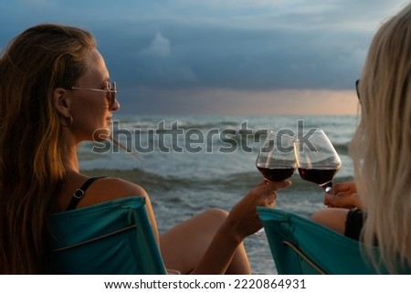 Two young beautiful women in swimsuits, sunglasses sitting on folding chairs on the beach clinking glasses with red wine at sunset, photo from behind