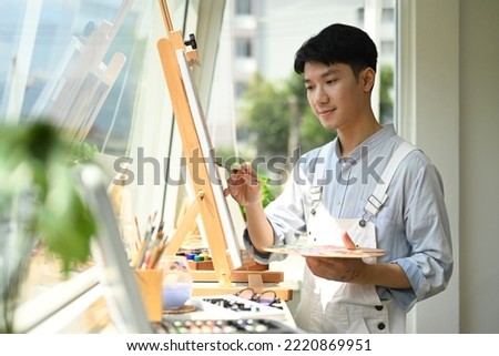 Concentrated asian man painting picture on canvas near window at art workshop. Leisure activity, creative hobby and art concept