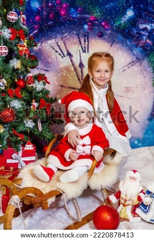 a small blue-eyed child and his sister in red Santa Claus costumes and red boots are sitting on real wooden sledges near the Christmas tree against the backdrop of a huge clock that shows 5 minutes to