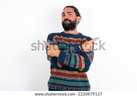 Serious young hispanic bearded man wearing knitted sweater over white background crosses hands and points at different sides hesitates between two items. Hard decision concept