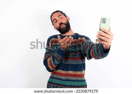 young hispanic bearded man wearing knitted sweater over white background blows air kiss at camera of smartphone and takes selfie, sends mwah via online call.