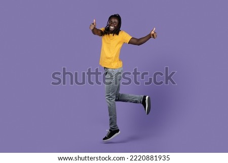 Joyful young black guy with long braids in stylish casual outfit jumping in the air on purple studio background, showing thumb ups and smiling, enjoying and recommending something, copy space