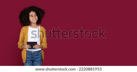 Happy preteen african american kid girl in casual with bushy hair using modern digital tablet, chatting with friends, looking at copy space over burgundy studio background, panorama