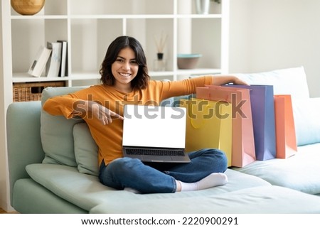Online Shopping. Happy Arab Woman Pointing At Laptop With Blank Screen While Sitting On Couch Next To Shopper Bags, Smiling Middle Eastern Lady Recommending Website For Internet Purchases, Mockup
