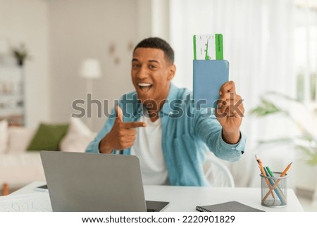 Cheerful excited millennial black guy with open mouth pointing finger to passport and air ticket, uses laptop in living room interior. End of covid-19 pandemic, travel and vacation, tourist recommends