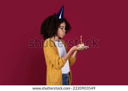 Adorable kid preteen girl with bushy hair having birthday party, wearing party hat, holding birthday cake, blowing candle, burgundy studio background, copy space. Birthday celebration