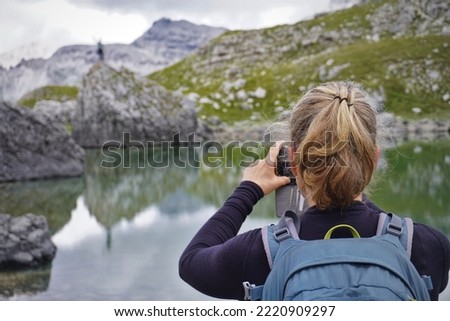 A woman taking photos with a phone of a alpine lake in mountain scenery 