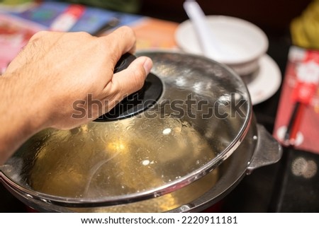 Action of people hand is preparing to open the Sukiyaki or Shabu Shabu pottery's lid after water already boiled. Selective focus photo. Royalty-Free Stock Photo #2220911181