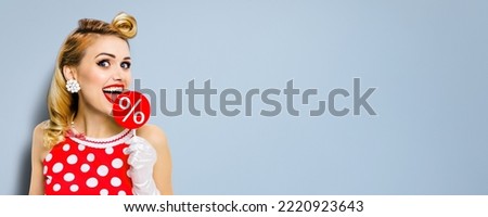 Very tasty discounts, rebates, deals, sales concept. Cheerful beautiful woman licking signboard with % sign, dressed in pinup red dress, isolated on grey background. Happy girl at studio. Wide picture