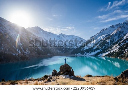 Far shot of a man standing on a huge boulder on a vantage point over stunning turquoise lake surrounded by snowy mountains on a sunny day. His hands are outstretched like wings, facing camera. Royalty-Free Stock Photo #2220936419