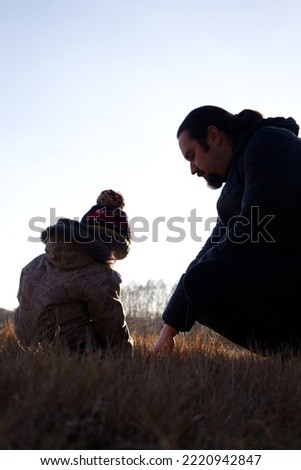 A little boy sits sits with his back to the camera next to his dad on the brown autumn grass in the field and examines something in the grass