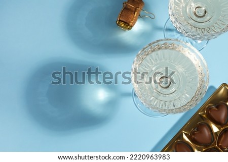 Glasses of expensive white wine, cork and heart shaped chocolate candies on light blue background, flat lay. Space for text