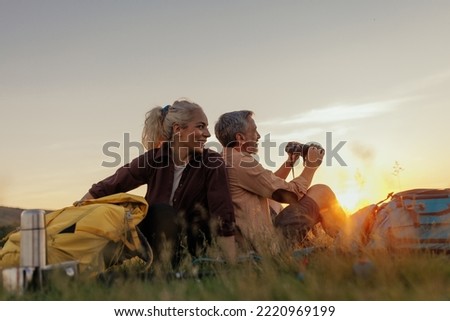 Middle age couple during their hike with backpacks. Sitting on meadow with their backs to each other and chilling Royalty-Free Stock Photo #2220969199