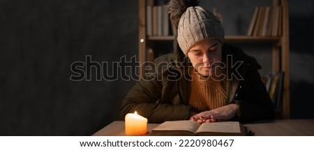 Young woman with warm clothing feeling the cold inside house, dark home no electricity, student studying using candlelight. Concept of no heating and no power in winter at home. Banner Royalty-Free Stock Photo #2220980467