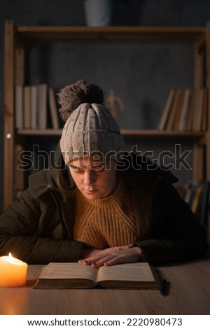 An angry student warmly dressed, feeling cold at home, studying by candlelight, a broken poor woman unable to pay for electricity. No heating, electric crisis in Europe Royalty-Free Stock Photo #2220980473