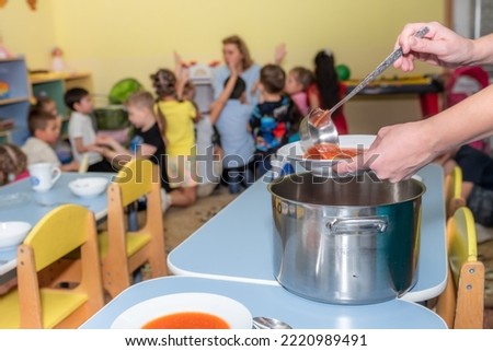 lunch in kindergarten. the nanny puts food on the children's plates. children play with the teacher. High quality photo Royalty-Free Stock Photo #2220989491