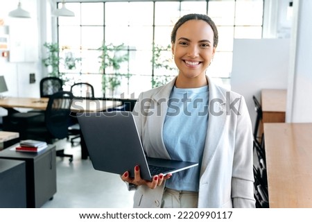 Gorgeous positive confident mixed race successful woman, seo, manager, stands in the office in elegant clothes, holds an open laptop in her hand, looks at camera, smiles friendly Royalty-Free Stock Photo #2220990117