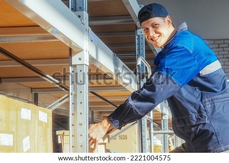 Storekeeper puts box on rack shelf looks at camera and smiles. Young Caucasian warehouse worker in overalls and baseball cap lays out goods inside warehouse. Authentic workflow. Royalty-Free Stock Photo #2221004575