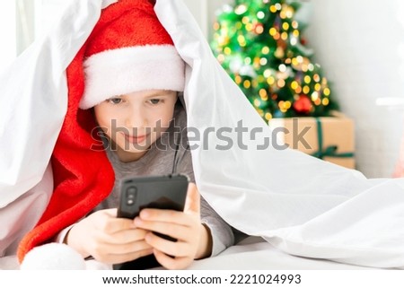 Child with a smartphone in his hands watches videos or cartoons in the bedroom. Boy is lying in bed and typing a congratulation message using the phone in front of the Christmas tree with gift boxes.