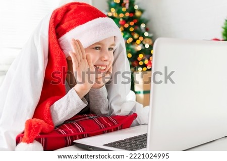 A smiling boy in a Santa hat congratulates his grandparents and friends on a video call. A joyful child waves his hand at the laptop screen while lying in front of the Christmas tree at home.
