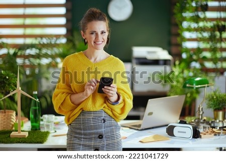 smiling modern middle aged small business owner woman in yellow sweater in the modern green office using smartphone.