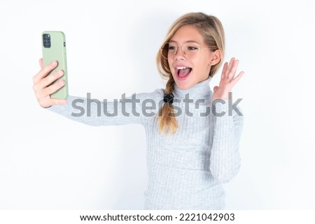 beautiful caucasian blond teen girl wearing gray turtleneck sweater over white wall holds modern mobile phone and makes video call waves palm in hello gesture. People modern technology concept