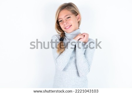 Charming serious beautiful caucasian teen girl wearing gray turtleneck sweater over white wall  keeps hands near face smiles tenderly at camera