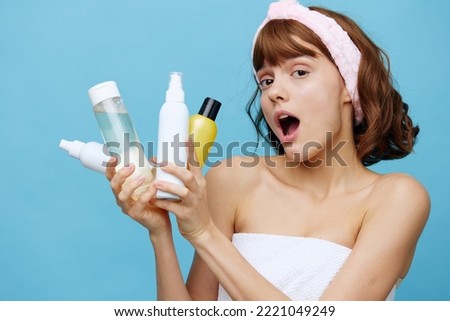 a shocked, surprised woman stands on a blue background, wrapped in a white towel and holding a set of jars with facial care cosmetics in her hands, her mouth wide open with emotions