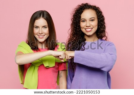 Young two friends smiling happy cheerful fun cool women 20s wearing green purple shirts looking camera giving fist bumb together isolated on pastel plain light pink color background studio portrait