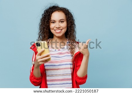 Young woman of African American ethnicity 20s she wear red jacket hold in hand use mobile cell phone show thumb up isolated on plain pastel light blue cyan background. Wet fall weather season concept