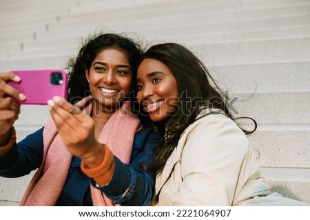Two young cheerful multinational women taking selfie photo while sitting on stairs outdoors