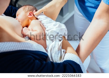 Unrecognizable midwife or breastfeeding consultant supporting a breastfeeding mother with her newborn baby girl in hospital ward. Proper attachment of the newborn during feeding. Royalty-Free Stock Photo #2221066183