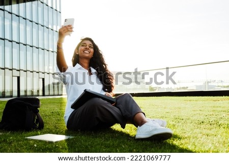 Young brunette indian woman using cellphone while sitting on grass outdoors
