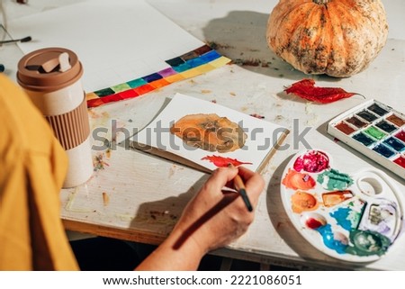 A woman paints a picture with a brush in a sketchbook. Learning to draw at home. Autumn still life with pumpkin and red leaf. Creating content concept.