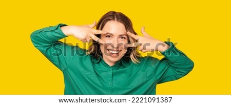 Playful blonde woman is showing v gesture with both hands near her face.