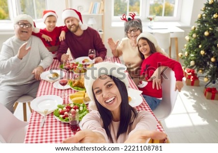 Happy mother taking selfie of whole family during festive dinner at home on Christmas day. Beautiful mom holding mobile phone, looking at camera and smiling together with rest of family in background