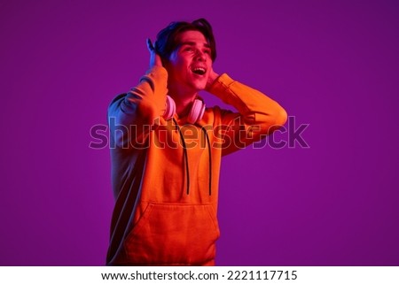 Portrait of young man in casual hoodie cheerfully posing with headphones isolated over purple background in neon light. Concept of youth, casual fashion, facial expression, emotions, lifestyle, ad